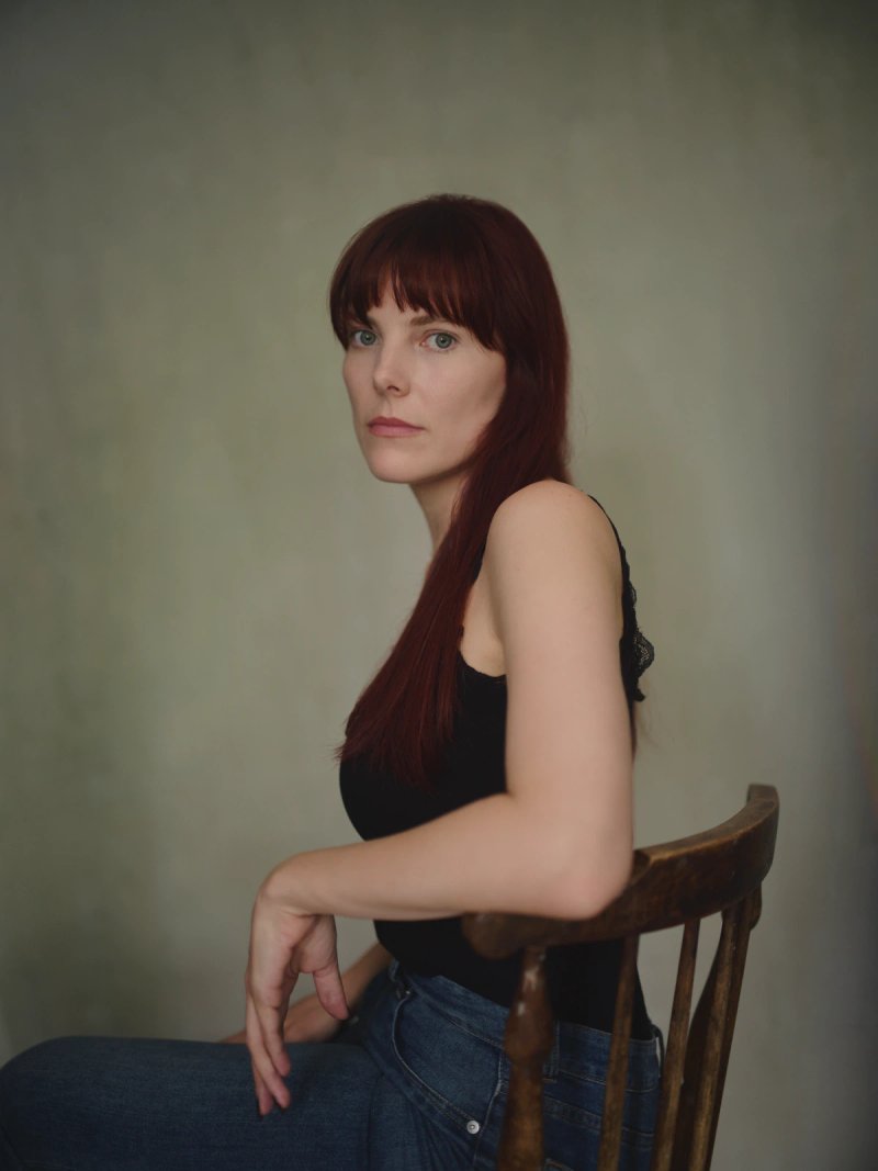 Actor portrait of Sophia seated on a wooden chair in my London studio, with long red hair, a black top and a grey-green canvas backdrop