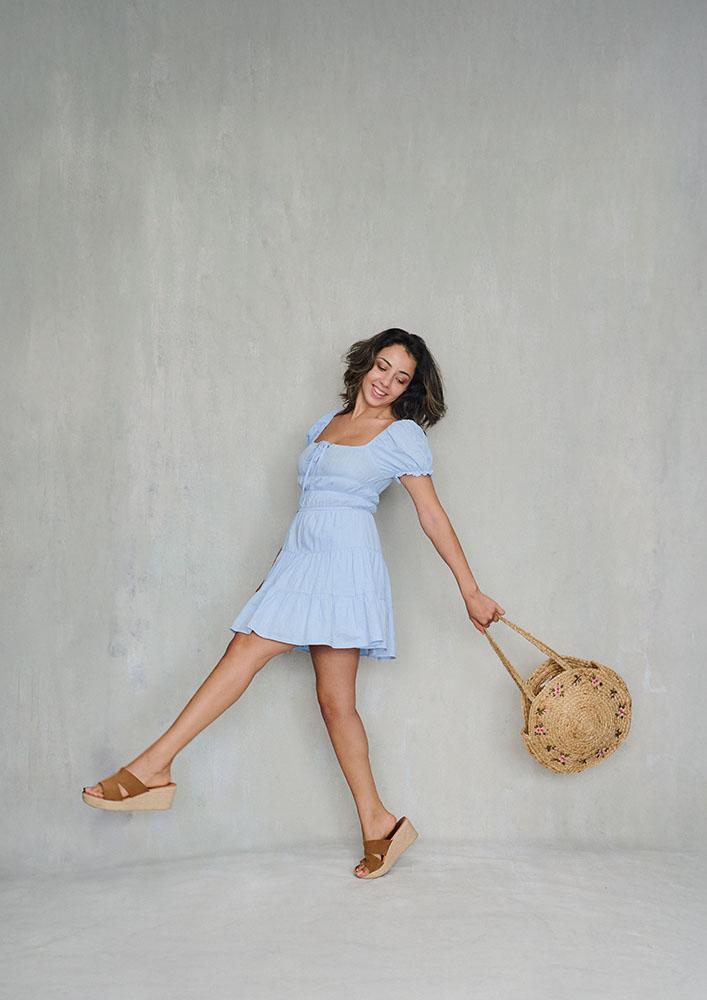 Portrait of female model with dark hair smiling and taking a large step whilst swinging a wicker bag.