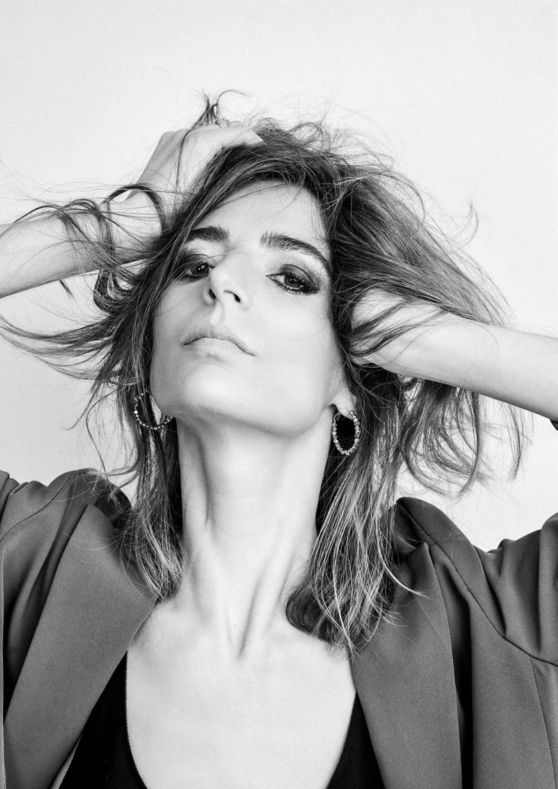 Black and white model portfolio portrait of a woman running her hands through tousled hair, wearing hoop earrings and an oversized blazer, against a white backdrop in my London studio.