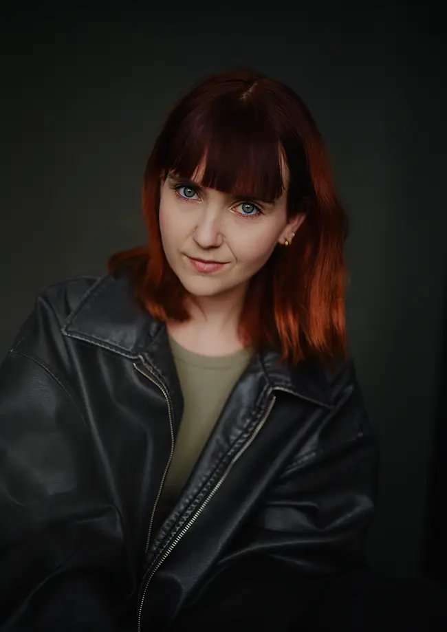Natural light seated portrait of actress Tess Hermann smiling in a London studio with a dark canvas backdrop