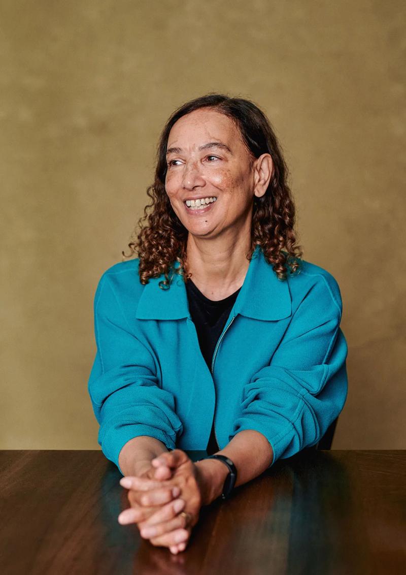 Author portrait of Jacqueline Crooks in a teal jacket, seated at a table against a painted canvas backdrop in my London studio.